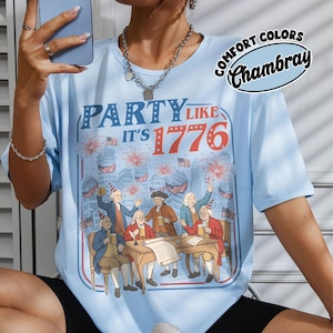 Puede incluir: Camiseta azul claro con el texto "PARTY LIKE IT'S 1776" y un gr&aacute;fico de figuras hist&oacute;ricas celebrando. La camiseta tambi&eacute;n presenta las palabras "COMFORT COLORS Chambray" en un dise&ntilde;o circular. El dise&ntilde;o incluye fuegos artificiales y banderas americanas.