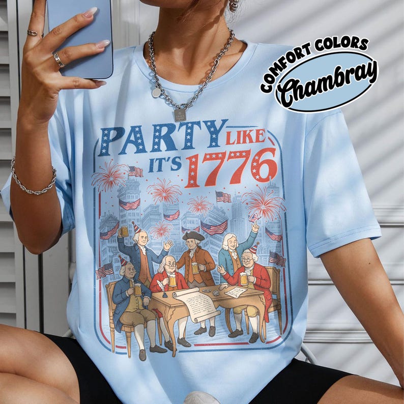Puede incluir: Camiseta azul claro con el texto "PARTY LIKE IT'S 1776" y un gr&aacute;fico de figuras hist&oacute;ricas celebrando. La camiseta tambi&eacute;n presenta las palabras "COMFORT COLORS Chambray" en un dise&ntilde;o circular. El dise&ntilde;o incluye fuegos artificiales y banderas americanas.