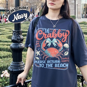 May include: Navy blue t-shirt with a crab graphic and the text "If Crabby, Please Return to the Beach." The design includes a crab, seashells, and starfish. The shirt is a Comfort Colors brand.