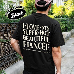 May include: Black t-shirt with the text "I LOVE MY SUPER-HOT BEAUTIFUL FIANC&Eacute;E so please stay away from me" in white. The shirt also has a "COMFORT COLORS Black" logo. The person is wearing a pink cap.