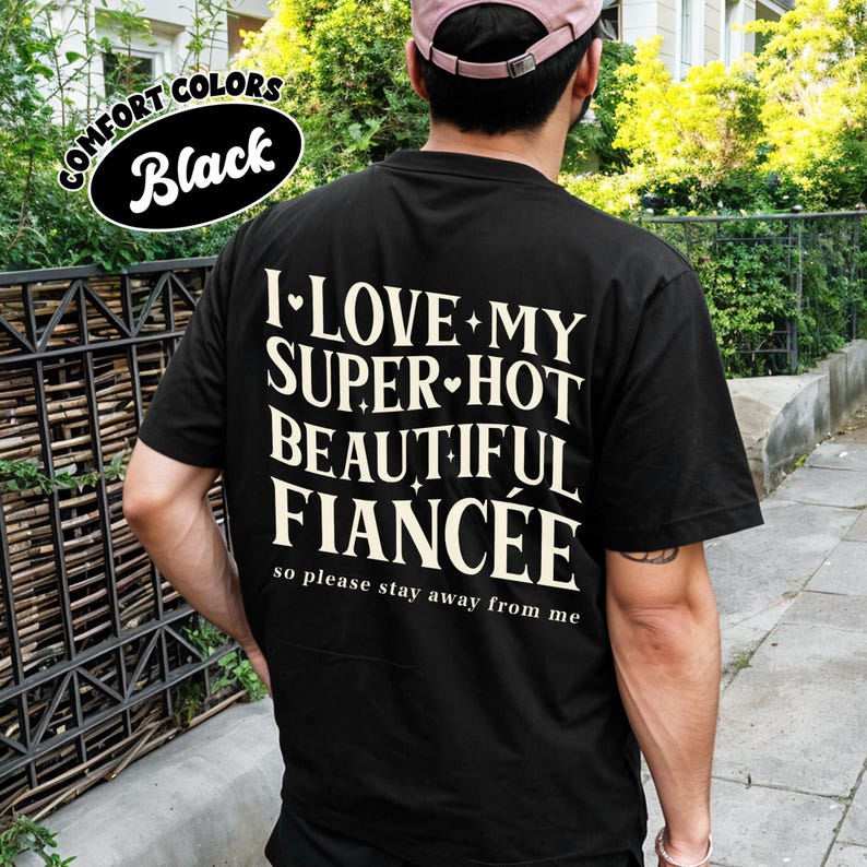 May include: Black t-shirt with the text "I LOVE MY SUPER-HOT BEAUTIFUL FIANC&Eacute;E so please stay away from me" in white. The shirt also has a "COMFORT COLORS Black" logo. The person is wearing a pink cap.