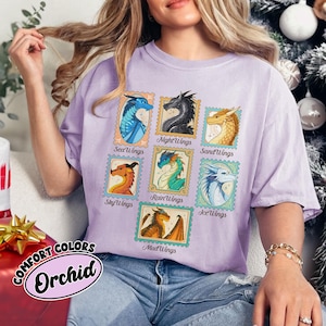 May include: A light purple t-shirt with a graphic of dragon stamps. The stamps feature different dragon designs with names like SeaWings, NightWings, and SandWings. The text "COMFORT COLORS Orchid" is printed on the bottom left.