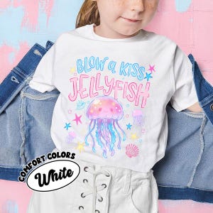 May include: White t-shirt with a colorful jellyfish graphic and the text "BLOW a KISS JELLYFISH". The design includes a pink jellyfish, stars, and a seashell. The shirt is labeled "COMFORT COLORS White".