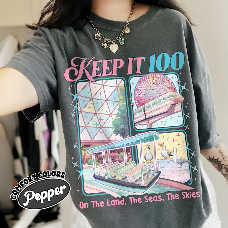 May include: Dark grey t-shirt with a colorful graphic featuring the text "KEEP IT 100" and illustrations of a futuristic sphere, a boat, and birds. The shirt also has the text "On The Land, The Seas, The Skies" and the brand name "Comfort Colors Pepper".