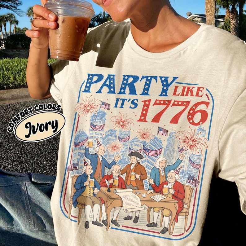 Puede incluir: Camiseta de color marfil con un gr&aacute;fico que dice "PARTY LIKE IT'S 1776" en azul y rojo. El gr&aacute;fico representa a un grupo de hombres con atuendo colonial celebrando con cerveza y fuegos artificiales. La camiseta tiene una etiqueta Comfort Colors.
