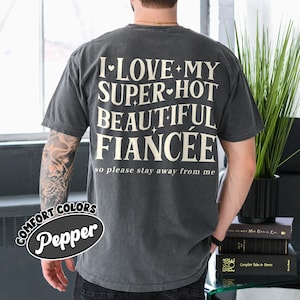 May include: A dark gray t-shirt with the text "I LOVE MY SUPER HOT BEAUTIFUL FIANC&Eacute;E so please stay away from me" in a white, stylized font. The shirt also has the words "COMFORT COLORS Pepper" in a black and white oval.