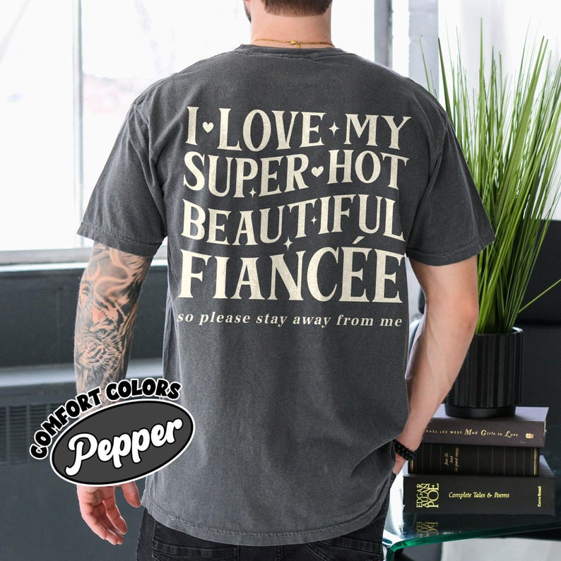 May include: A dark gray t-shirt with the text "I LOVE MY SUPER HOT BEAUTIFUL FIANC&Eacute;E so please stay away from me" in a white, stylized font. The shirt also has the words "COMFORT COLORS Pepper" in a black and white oval.