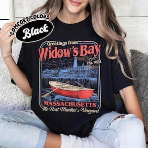 May include: A black t-shirt featuring a graphic print with the text Greetings from Widow's Bay, EST. 1681, MASSACHUSETTS, and The Next Martha's Vineyard. The design includes a red rowboat on water, a lighthouse, and buildings in a coastal scene.