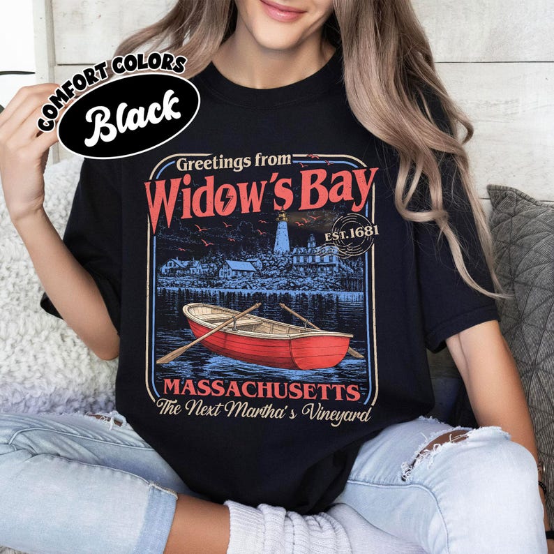 May include: A black t-shirt featuring a graphic print with the text Greetings from Widow's Bay, EST. 1681, MASSACHUSETTS, and The Next Martha's Vineyard. The design includes a red rowboat on water, a lighthouse, and buildings in a coastal scene.