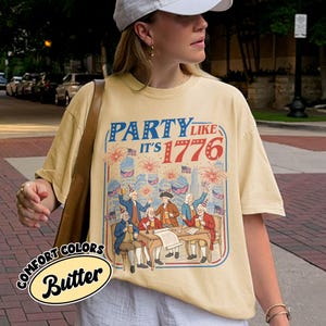 Puede incluir: Camiseta de color mantequilla con un gr&aacute;fico que dice "Party Like It's 1776". El dise&ntilde;o presenta ilustraciones de figuras hist&oacute;ricas, fuegos artificiales y banderas americanas. La camiseta tambi&eacute;n tiene las palabras "Comfort Colors" y "Butter".