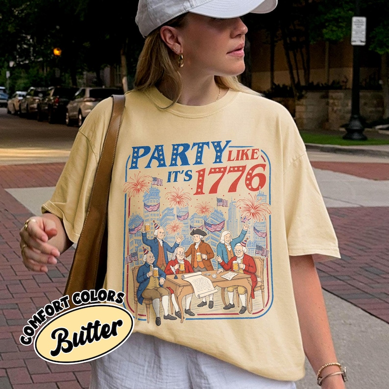 Puede incluir: Camiseta de color mantequilla con un gr&aacute;fico que dice "Party Like It's 1776". El dise&ntilde;o presenta ilustraciones de figuras hist&oacute;ricas, fuegos artificiales y banderas americanas. La camiseta tambi&eacute;n tiene las palabras "Comfort Colors" y "Butter".