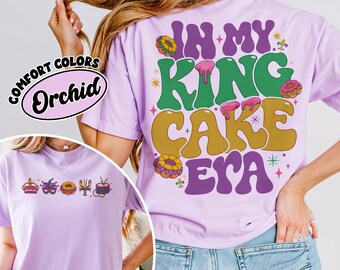May include: A purple t-shirt with the text "In My King Cake Era" in a retro font. The text is surrounded by images of king cakes and Mardi Gras decorations. The shirt is available in a comfort color called "Orchid".