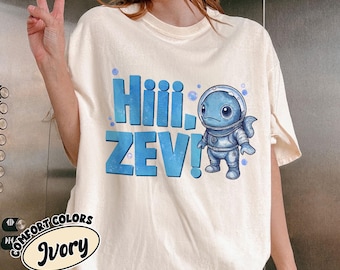 May include: Ivory-colored t-shirt with the text "Hiii, ZEV!" in blue. A blue cartoon astronaut character is to the right of the text. The shirt is part of the Comfort Colors collection.