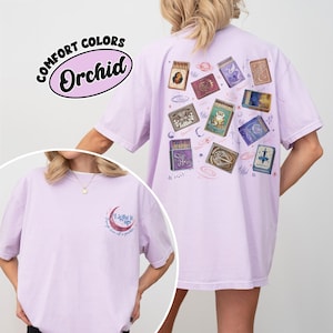 May include: A light purple t-shirt with a graphic of matchboxes on the back. The matchboxes have various designs, including celestial and animal themes. The front of the shirt has a crescent moon graphic with the text "Light it up". The shirt is labeled "Comfort Colors Orchid".