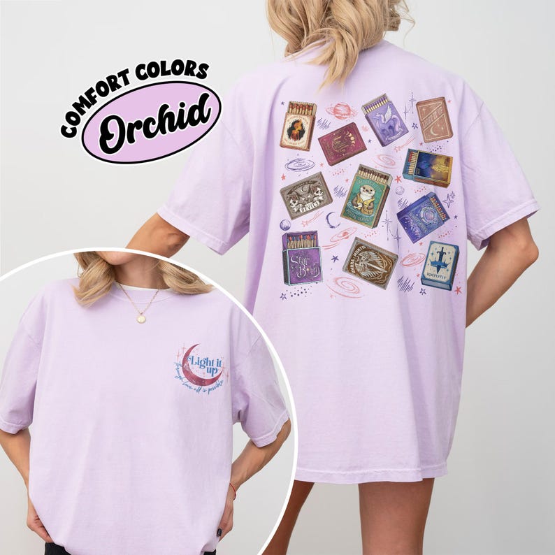 May include: A light purple t-shirt with a graphic of matchboxes on the back. The matchboxes have various designs, including celestial and animal themes. The front of the shirt has a crescent moon graphic with the text "Light it up". The shirt is labeled "Comfort Colors Orchid".
