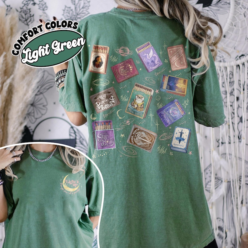 May include: Light green t-shirt with a collection of matchbox illustrations on the back. The matchboxes feature various designs, including a unicorn, a frog, and celestial elements. The shirt also has the text "Comfort Colors" and "Light Green".