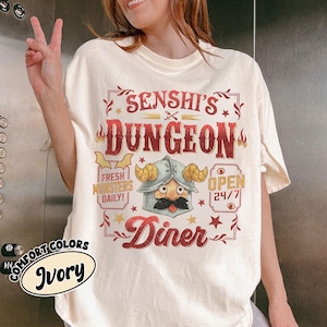 May include: Ivory-colored t-shirt with a graphic design featuring the text "SENSHI'S DUNGEON Diner" in red and a cartoon character with a mustache and helmet. The shirt also has the text "FRESH MONSTERS DAILY!" and "OPEN 24/7".