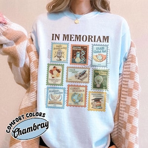 May include: Light blue sweatshirt with the words "IN MEMORIAM" above a collection of postage stamp designs. The stamps feature illustrations and text related to various topics, including "Public Broadcasting" and "Climate Research". The sweatshirt also has the words "COMFORT COLORS Chambray".