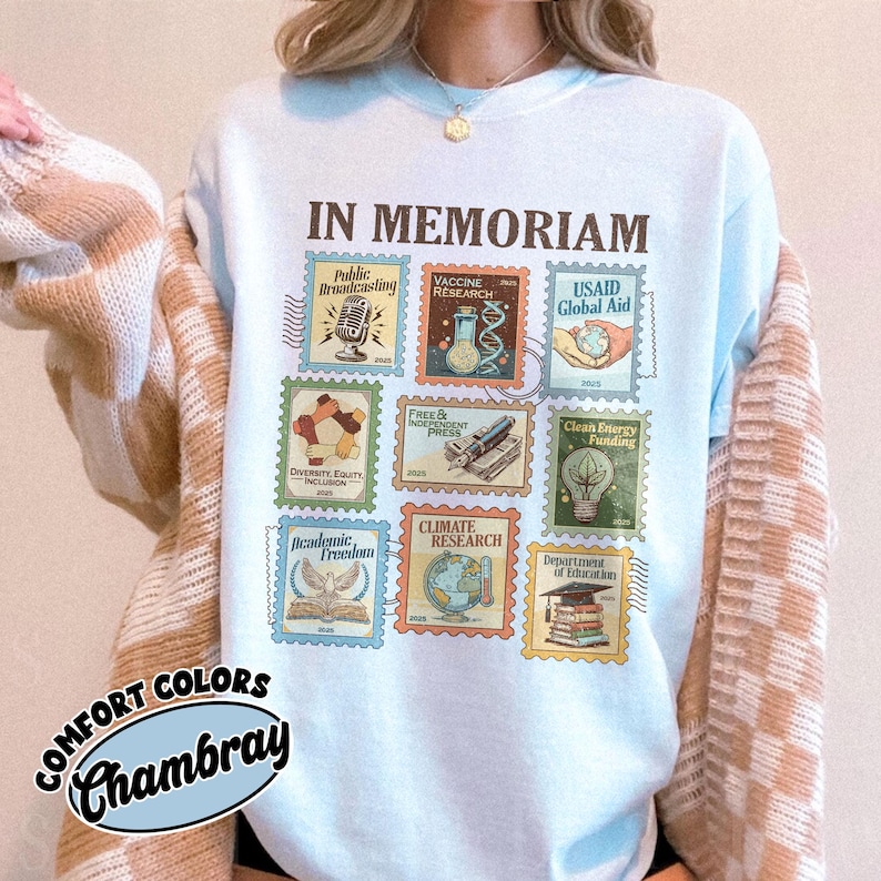 May include: Light blue sweatshirt with the words "IN MEMORIAM" above a collection of postage stamp designs. The stamps feature illustrations and text related to various topics, including "Public Broadcasting" and "Climate Research". The sweatshirt also has the words "COMFORT COLORS Chambray".