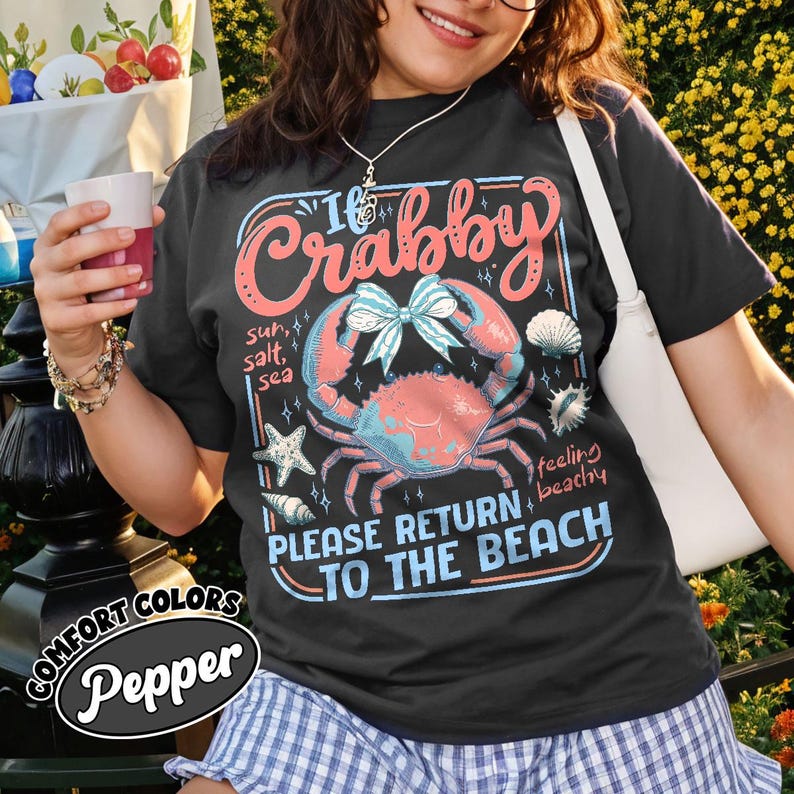 May include: Black t-shirt with a crab graphic and the text "I'm Crabby, Please Return to the Beach." The design includes a crab, seashells, and starfish in shades of pink, blue, and white. The shirt is from Comfort Colors.
