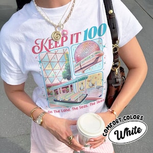 May include: White t-shirt with a colorful graphic design featuring the text "KEEP IT 100" and illustrations of a boat, train, and geometric shapes. The shirt also has the text "On The Land. The Seas. The Skies." and "COMFORT COLORS White".