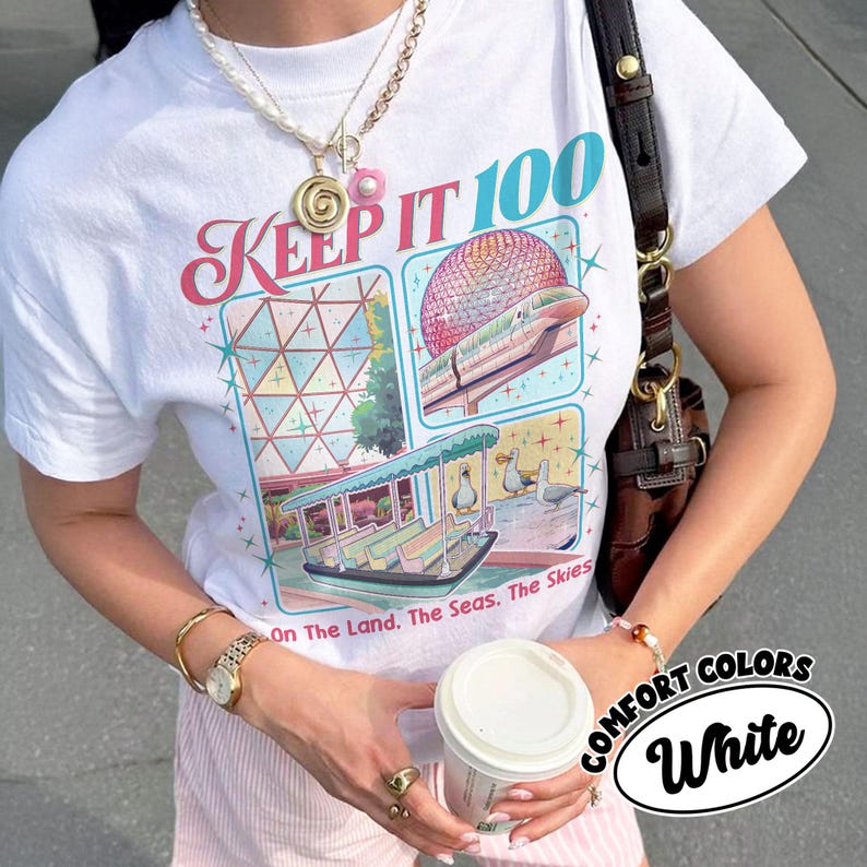 May include: White t-shirt with a colorful graphic design featuring the text "KEEP IT 100" and illustrations of a boat, train, and geometric shapes. The shirt also has the text "On The Land. The Seas. The Skies." and "COMFORT COLORS White".