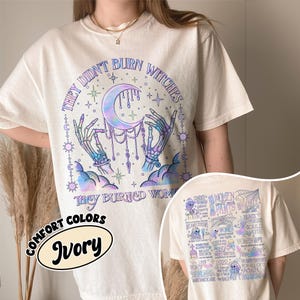 May include: Ivory-colored t-shirt with a graphic on the front featuring a crescent moon, skeletal hands, and the text "THEY DIDN'T BURN WITCHES THEY BURNED WOMEN." The back of the shirt has a graphic with text and illustrations.