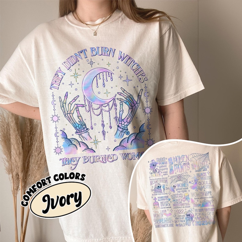 May include: Ivory-colored t-shirt with a graphic on the front featuring a crescent moon, skeletal hands, and the text "THEY DIDN'T BURN WITCHES THEY BURNED WOMEN." The back of the shirt has a graphic with text and illustrations.