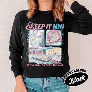 May include: Black long-sleeve shirt with the text "KEEP IT 100" in pink and blue. The shirt features four illustrated squares with a boat, a building, a monorail, and ducks. The text "On The Land. The Seas. The Skies" is below the squares. The shirt is labeled "COMFORT COLORS Black".