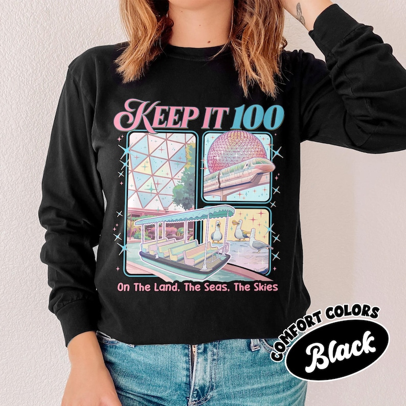May include: Black long-sleeve shirt with the text "KEEP IT 100" in pink and blue. The shirt features four illustrated squares with a boat, a building, a monorail, and ducks. The text "On The Land. The Seas. The Skies" is below the squares. The shirt is labeled "COMFORT COLORS Black".