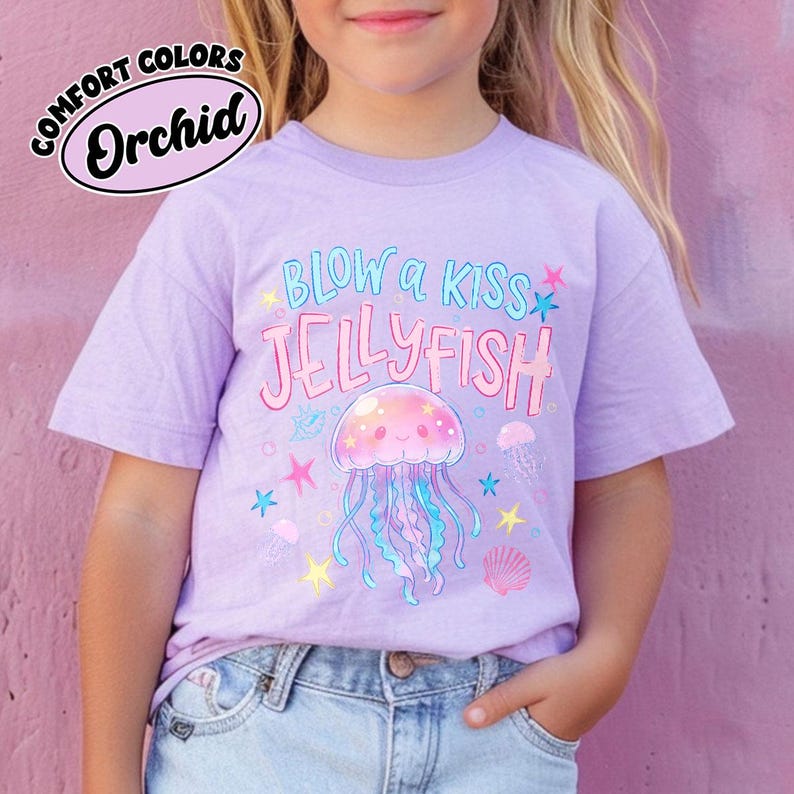 May include: A light purple t-shirt with the words "BLOW a KISS JELLYFISH" in a playful font, featuring a cartoon jellyfish illustration. The shirt also has the words "COMFORT COLORS" and "Orchid" in a circular design.