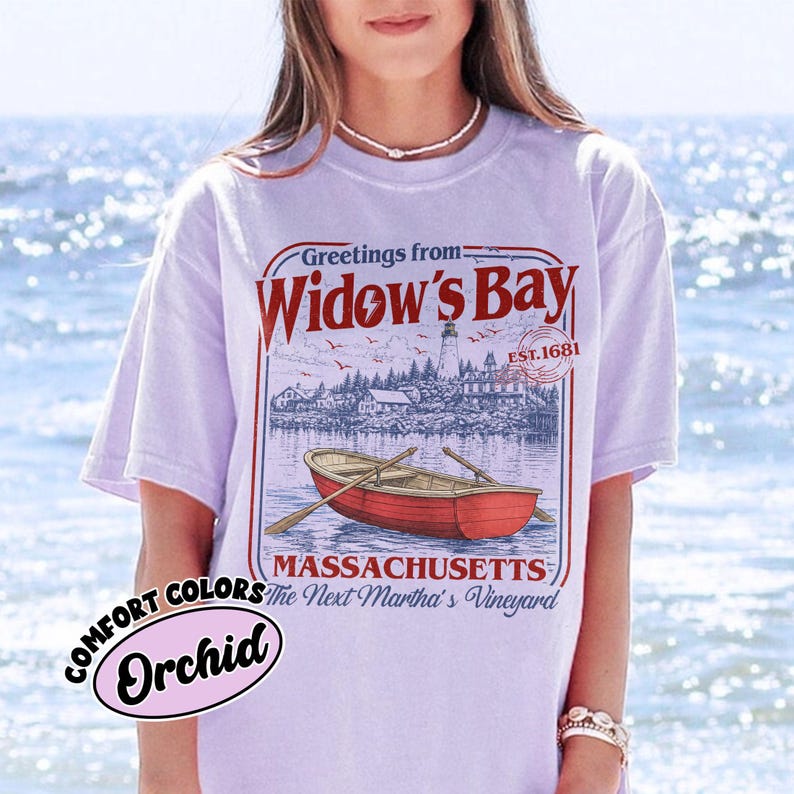 May include: A light purple short-sleeve t-shirt featuring a graphic print with the text Greetings from Widow's Bay, EST. 1681, Massachusetts, The Next Martha's Vineyard, a lighthouse, and a red rowboat with oars on the water.