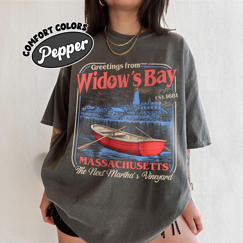 May include: A charcoal gray t-shirt featuring a graphic print with the text Greetings from Widow's Bay, Est. 1681, Massachusetts, The Next Martha's Vineyard. The design includes a red rowboat on water and a lighthouse in the background.
