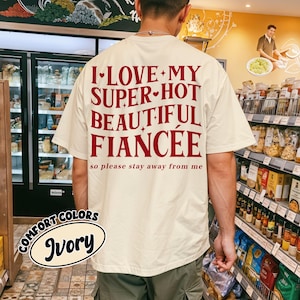 May include: Ivory-colored t-shirt with the text "I LOVE MY SUPER-HOT BEAUTIFUL FIANC&Eacute;E so please stay away from me" in red. The shirt is from Comfort Colors. The person is standing in a grocery store.