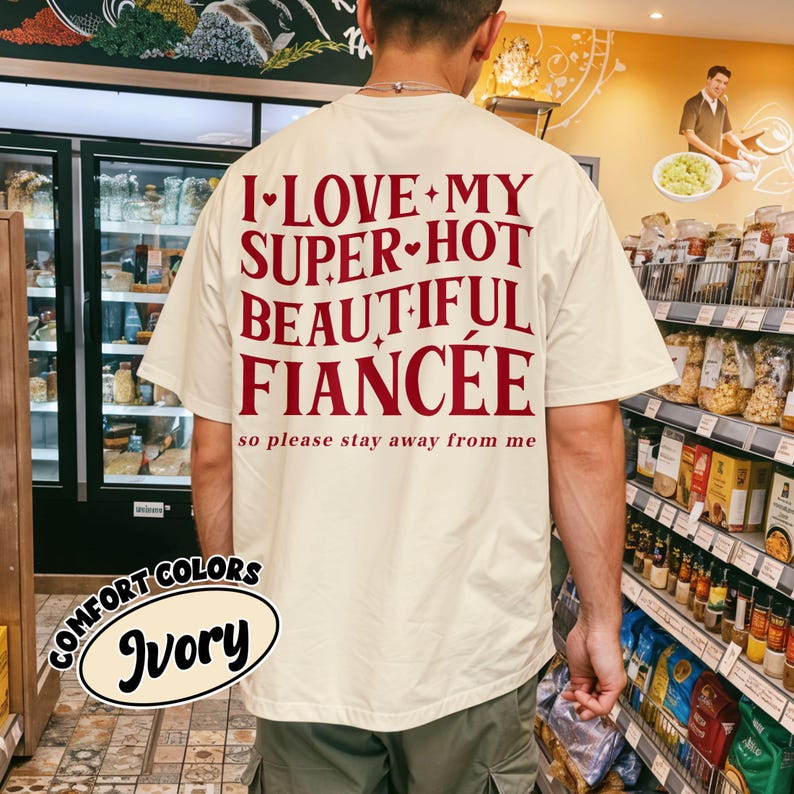 May include: Ivory-colored t-shirt with the text "I LOVE MY SUPER-HOT BEAUTIFUL FIANC&Eacute;E so please stay away from me" in red. The shirt is from Comfort Colors. The person is standing in a grocery store.