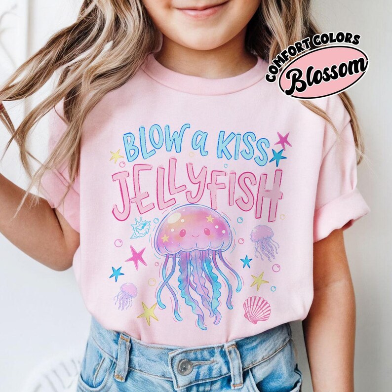 May include: A light pink t-shirt with the text "BLOW a KISS JELLYFISH" in blue and pink. The shirt features a cartoon jellyfish with blue and purple tentacles, surrounded by stars and seashells. The shirt is labeled "COMFORT COLORS Blossom".