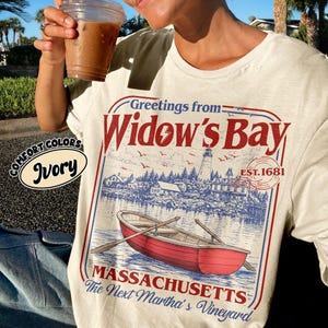 May include: An ivory t-shirt featuring a graphic print with the text Greetings from Widow's Bay, Est. 1681, Massachusetts, The Next Martha's Vineyard. The design includes a red rowboat on water, a lighthouse, and coastal buildings in blue.