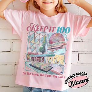 May include: Pink t-shirt with the text "KEEP IT 100" and illustrations of a boat, train, and sphere. The shirt also has the text "On The Land, The Seas, The Skies" and the words "COMFORT COLORS Blossom" in a circle.