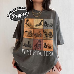 May include: Dark grey t-shirt with a grid of monkey illustrations in various poses. The shirt features the text "IN MY PUNCH ERA" in white, with a "Comfort Colors Pepper" logo at the top. The shirt has short sleeves and a relaxed fit.