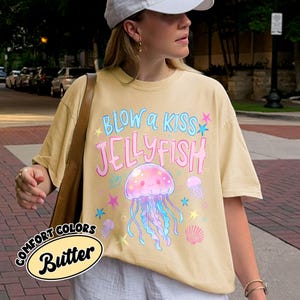 May include: A light yellow t-shirt with the text "BLOW a KISS JELLYFISH" in blue and pink. The shirt features a cartoon jellyfish with blue and pink tentacles, surrounded by stars and other jellyfish. The shirt also has the words "COMFORT COLORS Butter" in a circular design.