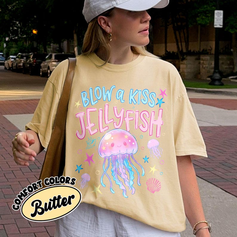 May include: A light yellow t-shirt with the text "BLOW a KISS JELLYFISH" in blue and pink. The shirt features a cartoon jellyfish with blue and pink tentacles, surrounded by stars and other jellyfish. The shirt also has the words "COMFORT COLORS Butter" in a circular design.