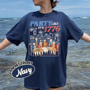 Puede incluir: Camiseta azul marino con la frase "PARTY LIKE IT'S 1776". El dise&ntilde;o muestra un grupo de personas alrededor de una mesa, con fuegos artificiales y un horizonte urbano de fondo. Tambi&eacute;n aparece el texto "Comfort Colors Navy".