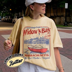 May include: A butter yellow short-sleeve t-shirt featuring a graphic print with the text Greetings from Widow's Bay, EST. 1681, Massachusetts, The Next Martha's Vineyard. The design includes a red rowboat, a lighthouse, and coastal scenery.