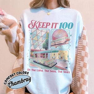 May include: Light blue Comfort Colors Chambray sweatshirt with the text "KEEP IT 100" and illustrations of a monorail, boat, and other theme park elements. The text "On The Land. The Seas. The Skies" is also present.
