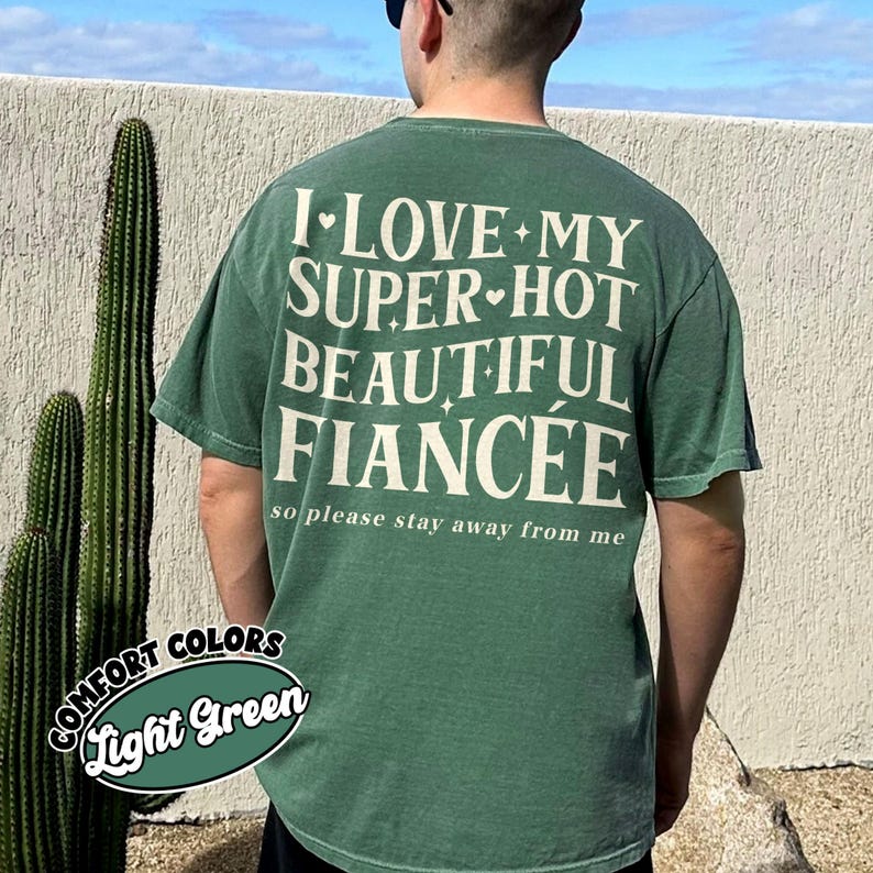 May include: Light green t-shirt with the text "I LOVE MY SUPER HOT BEAUTIFUL FIANC&Eacute;E so please stay away from me" in white. The shirt also has a "COMFORT COLORS Light Green" label. A person is wearing the shirt.