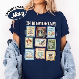 May include: Navy blue t-shirt with a graphic of postage stamps. The stamps feature images and text including "IN MEMORIAM", "Public Broadcasting", "Vaccine Research", and "USAID Global Aid". The shirt also has the words "COMFORT COLORS" and "Navy" in a circular design.