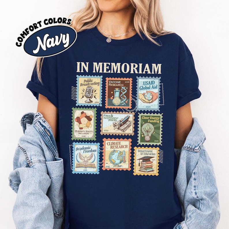 May include: Navy blue t-shirt with a graphic of postage stamps. The stamps feature images and text including "IN MEMORIAM", "Public Broadcasting", "Vaccine Research", and "USAID Global Aid". The shirt also has the words "COMFORT COLORS" and "Navy" in a circular design.