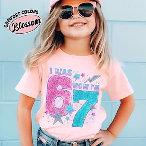 May include: A light pink t-shirt with the text "I WAS 6 NOW I'M 7" in a colorful design. The number 6 is pink and the number 7 is teal. The shirt also features stars and lightning bolts.