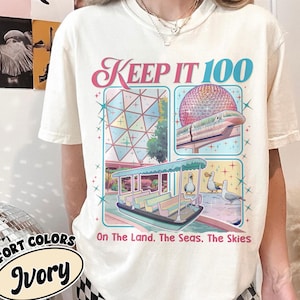 May include: Ivory-colored t-shirt with the text "KEEP IT 100" in pink and blue. The shirt features illustrations of a futuristic building, a monorail, and a boat with ducks. The text "On The Land, The Seas, The Skies" is also included.