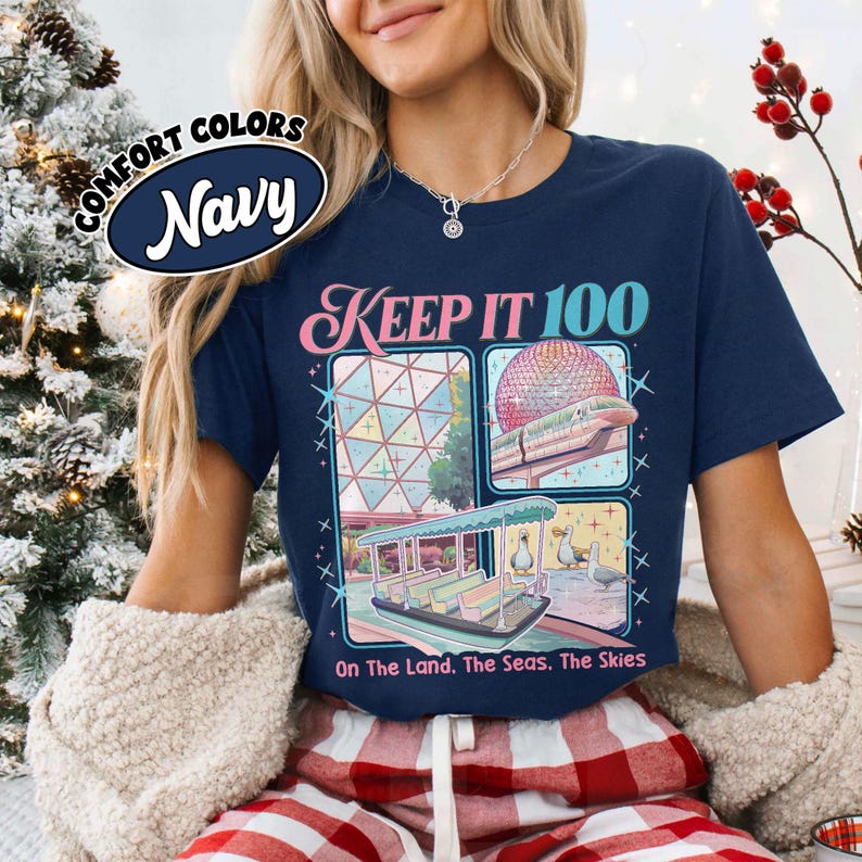 May include: Navy blue t-shirt with the text "KEEP IT 100" and three illustrated panels. The panels depict a geometric structure, a monorail with a geodesic sphere, and a boat with ducks. The shirt also has the text "On The Land. The Seas. The Skies."